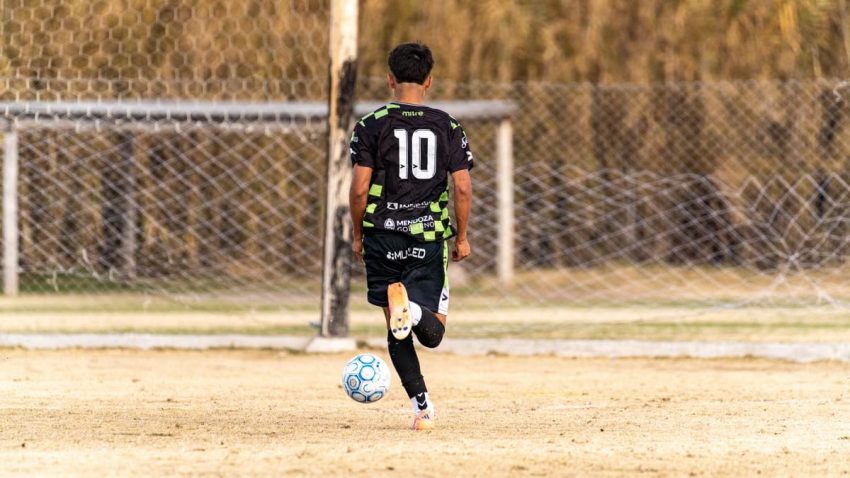 Teen soccer player dribbles the ball towards the goal on a dusty field.